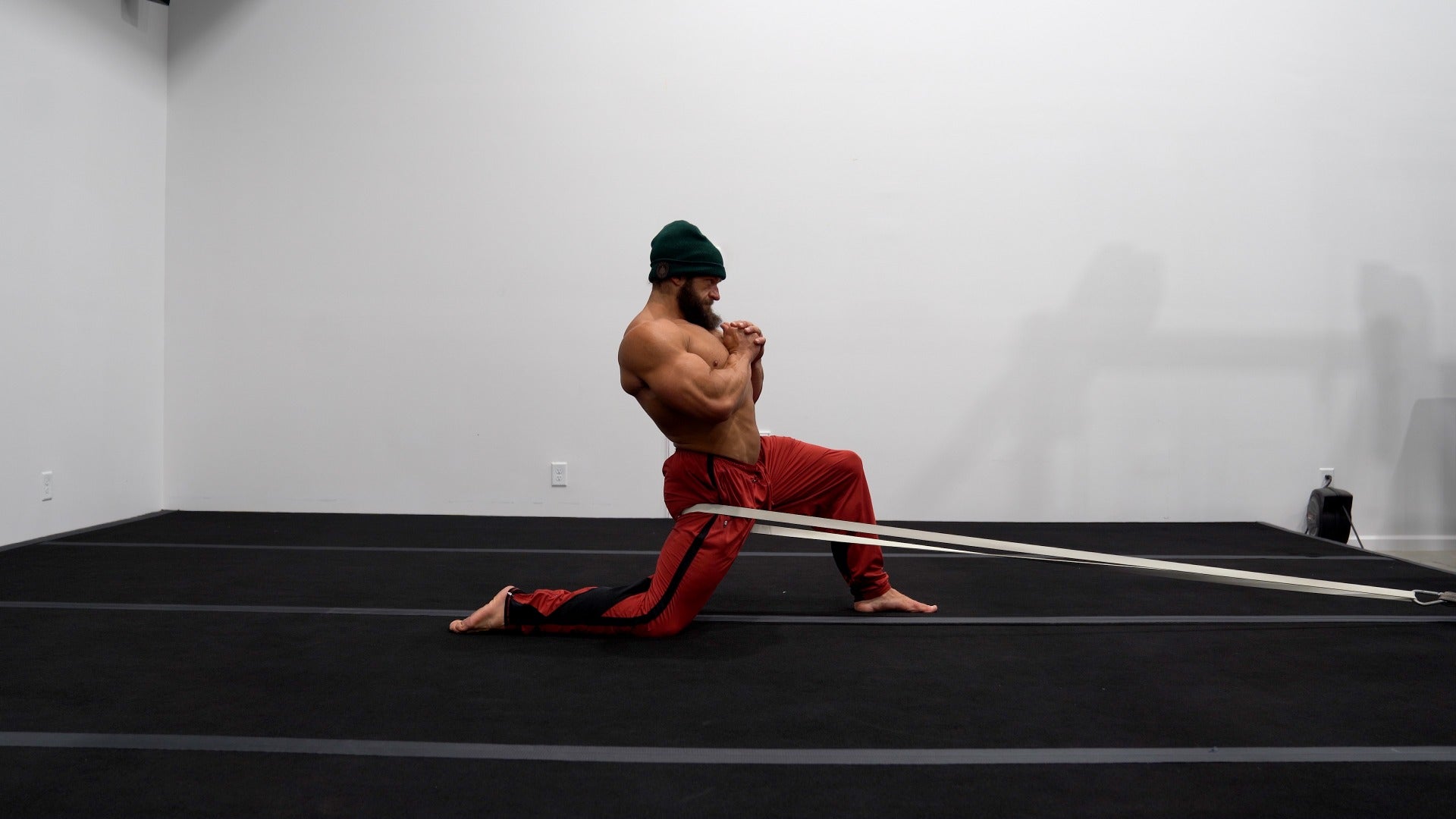 Person in red pants and green headband performing a martial arts move with a white belt on a black mat.